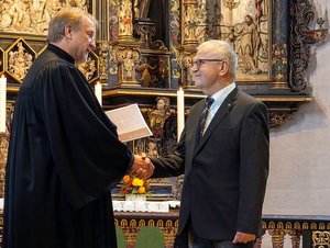 Ein Mann ist geistlicher Kleidung überreicht einem älteren Mann im Anzug eine Urkunde. Im Hintergrund ist ein Altar zu sehen.